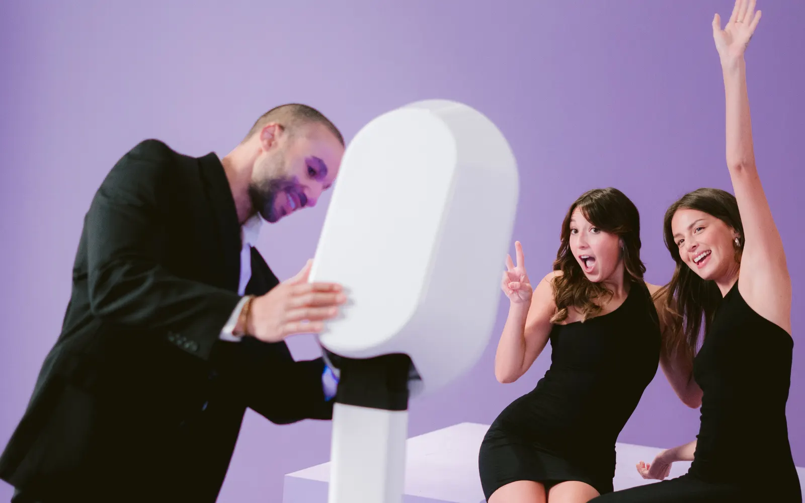 A man in a dark suit leaning into an oval photo booth while two women in black dresses pose and cheer beside him against a lavender wall.