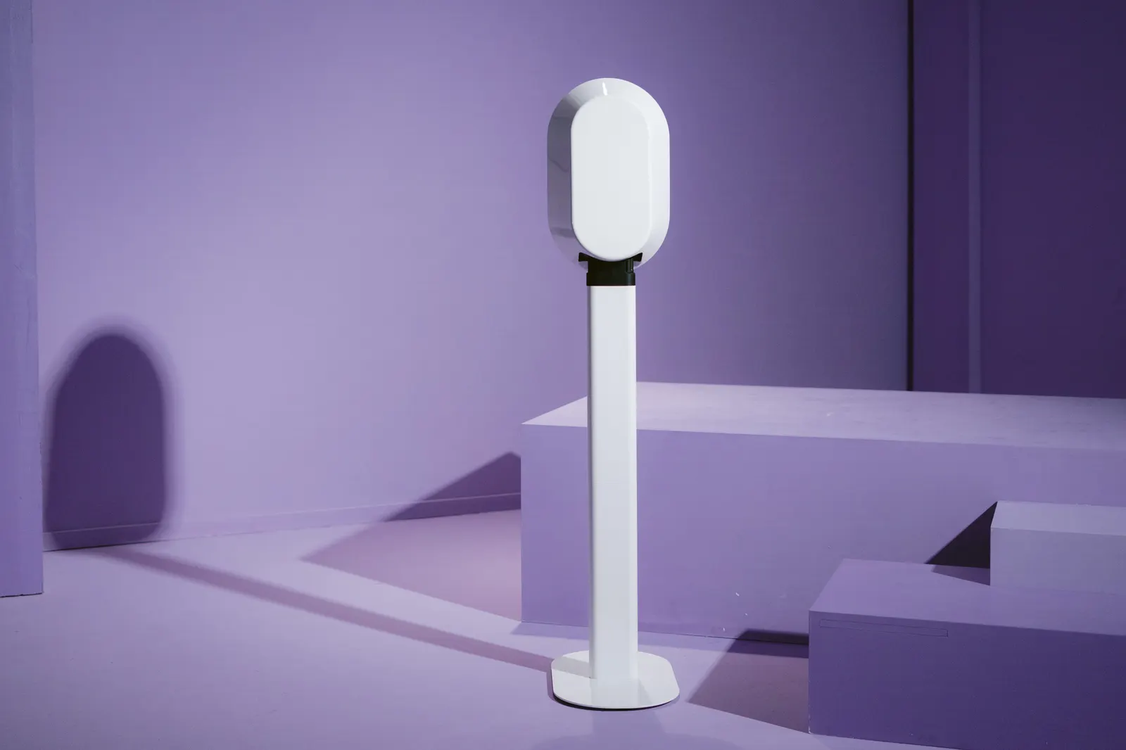 A white oval open-air photo booth on a slim stand against a lavender backdrop with soft blocks of seating behind it.