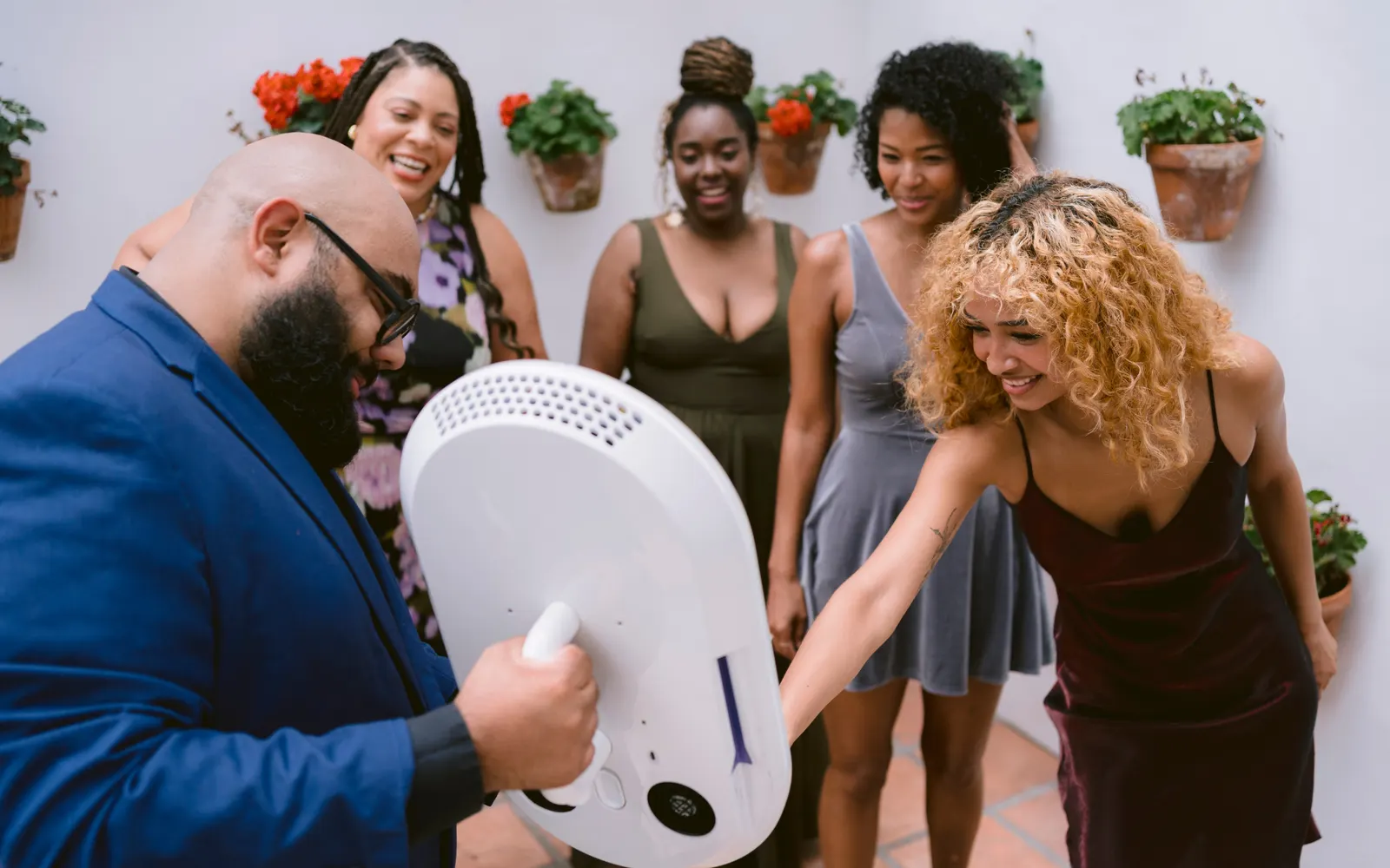Guests crowding around a white photo booth head in a whitewashed courtyard, one man holding the handle while friends reach in to pose.