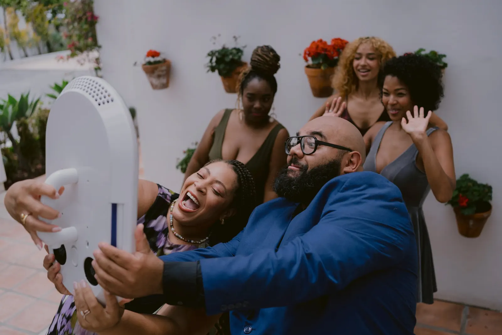 A group of friends squeezing in together and laughing as they take photos with a handheld photo booth head in a courtyard.